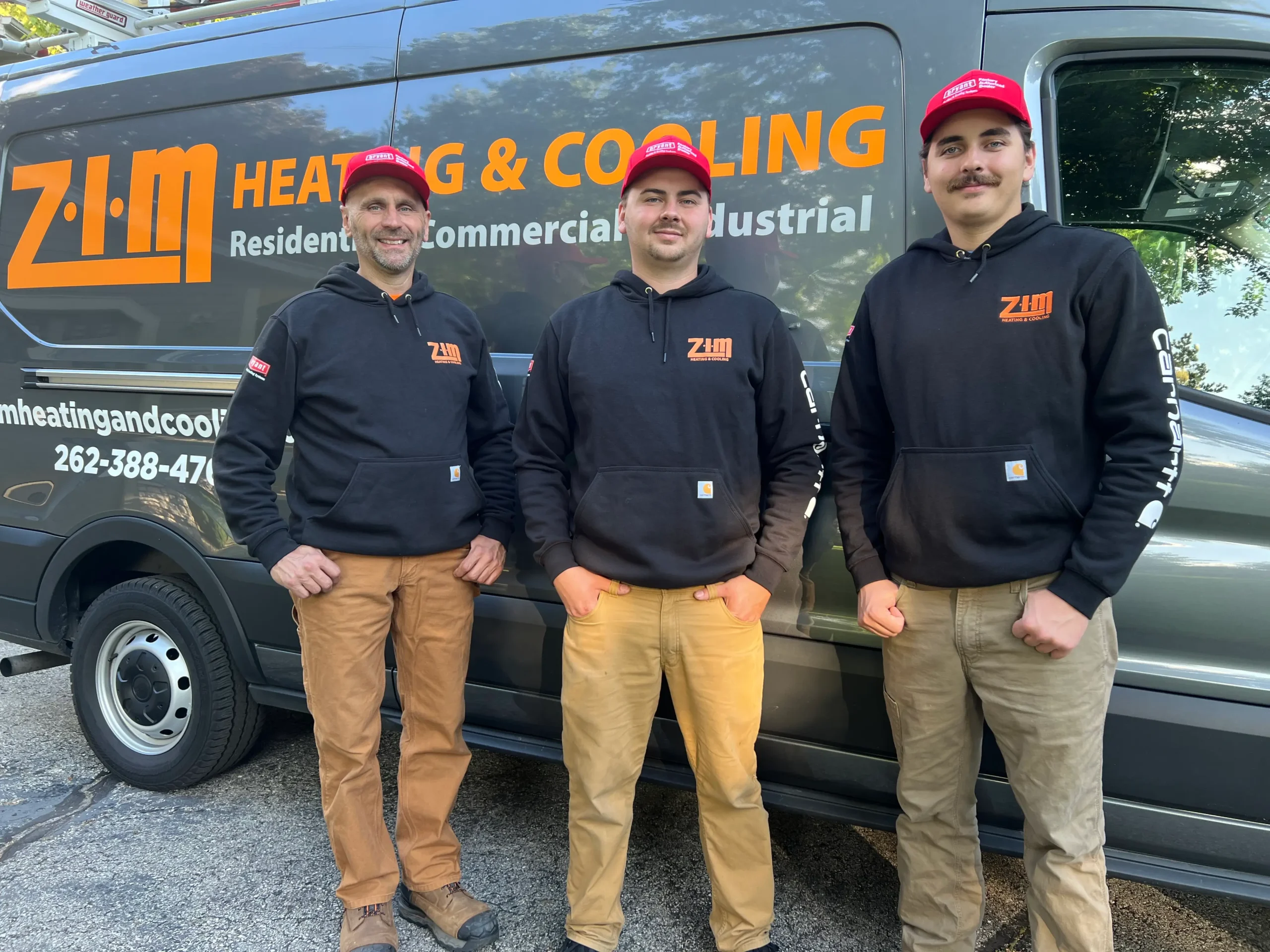 Three technicians for ZIM Heating and Cooling HVAC Services standing in front of company van in Waukesha, WI wearing black company sweatshirts and red hats